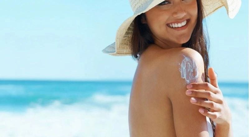 Mulher na praia de chapéu passando protetor no ombro