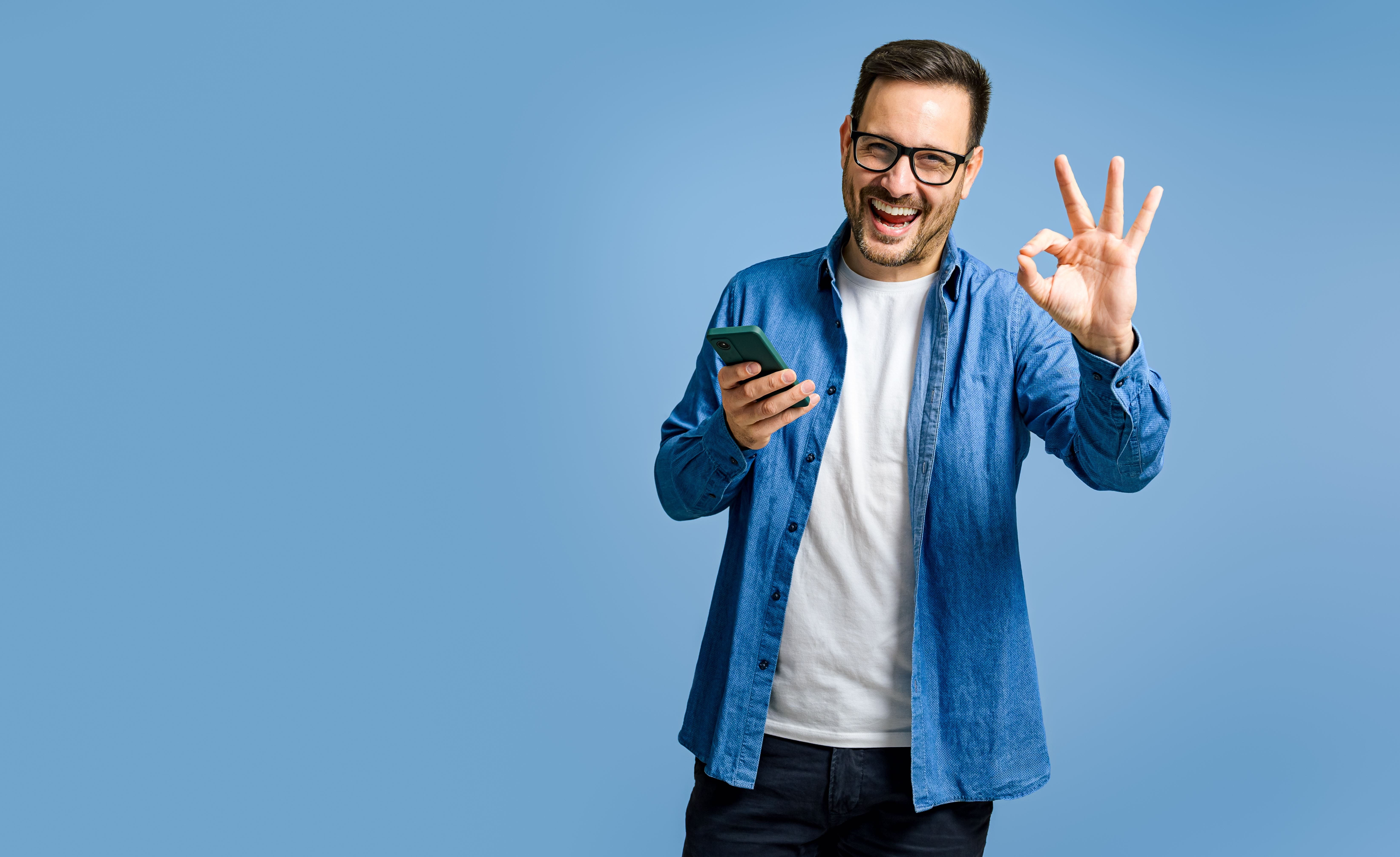 Mann im blauen Shirt hält ein Smartphone und macht eine „OK“-Geste.
