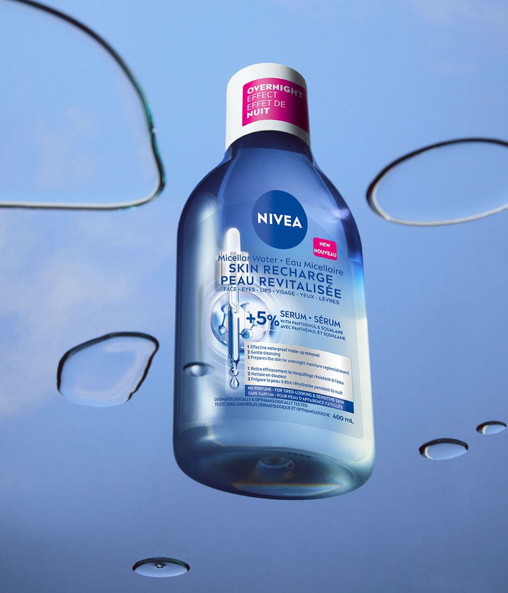 Un flacon d'Eau Micellaire NIVEA Peau revitalisée infusée au sérum est présenté sur un arrière-plan bleu, entouré de gouttelettes d'eau dispersées. L'étiquette du flacon précise l'effet de nuit du produit et sa formule à 5 % de sérum avec panthénol et squalane.