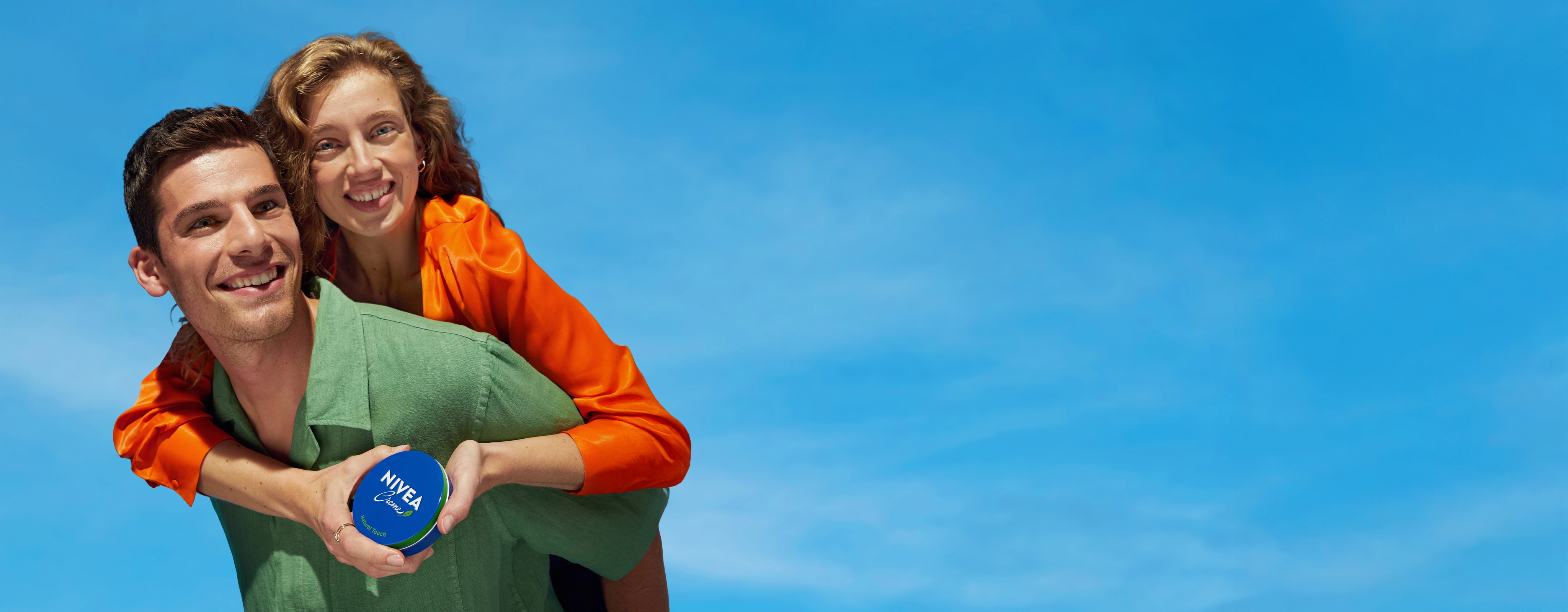 Smiling woman holding NIVEA Creme rides on man’s back under a clear blue sky, both joyful.