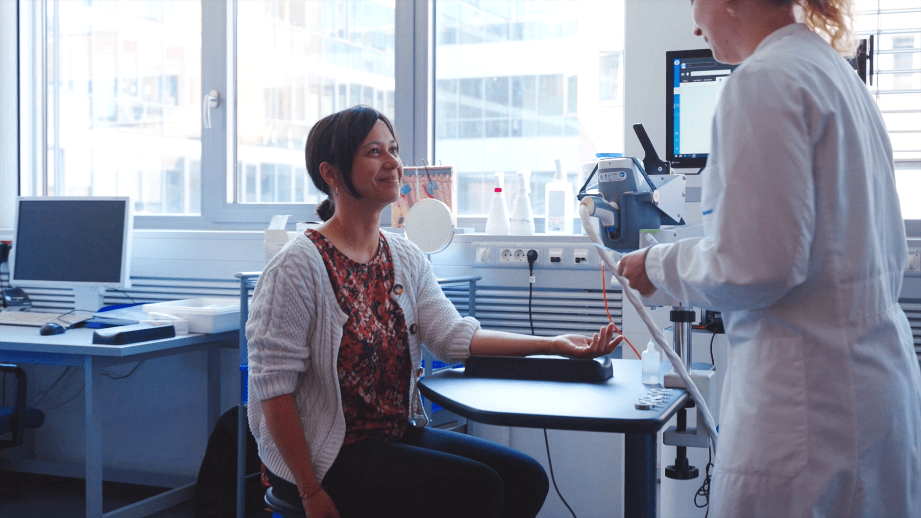 A woman in a lab coat assists a seated woman in a medical setting.