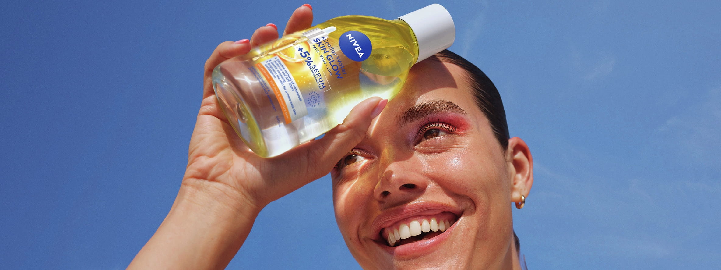 Kvinde smiler og holder NIVEA Skin Glow Serum-Infused Micellar Water mod panden, der kaster skygger