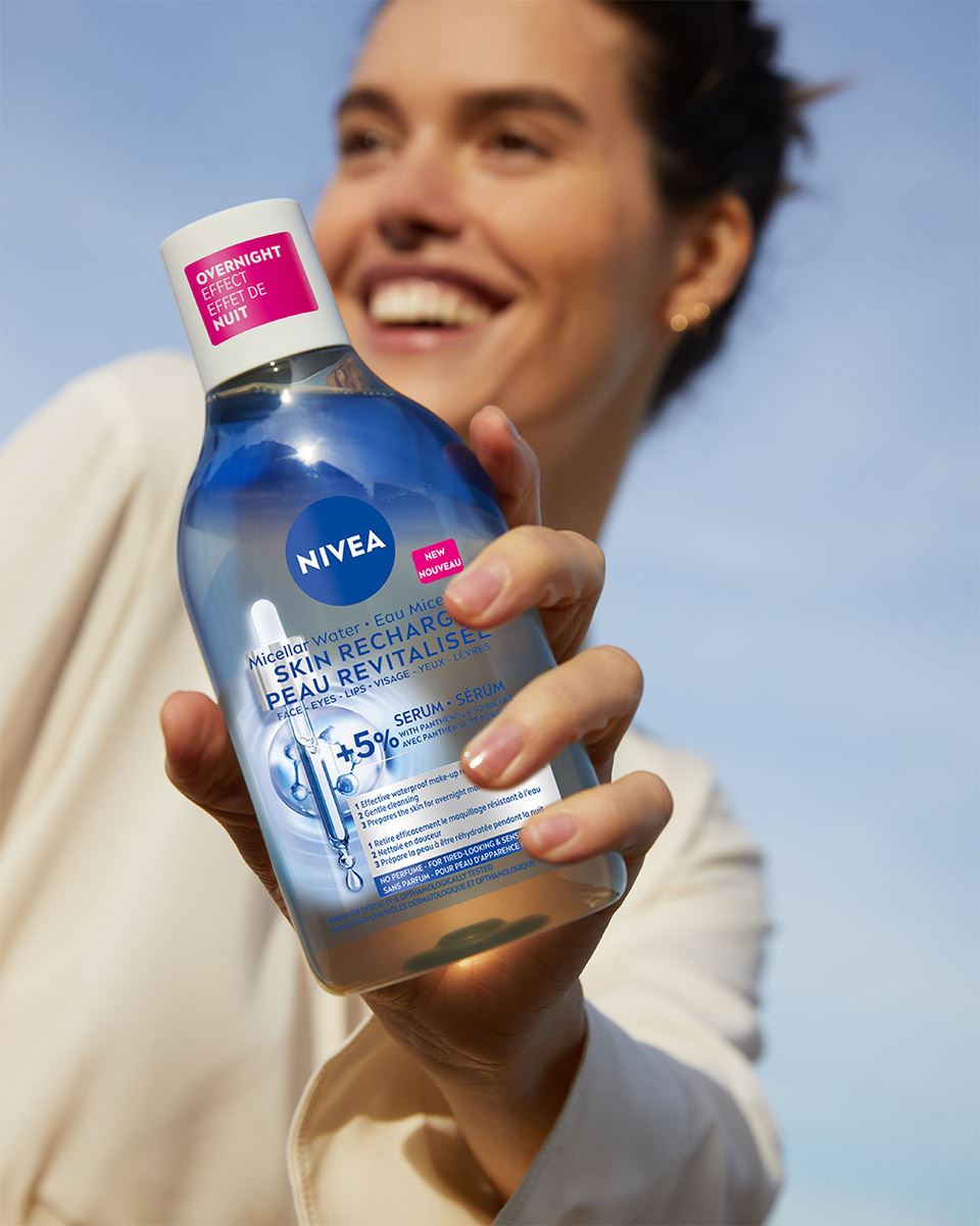 A woman smiling while holding a bottle of NIVEA Skin Recharge Serum-Infused Micellar Water. The bottle features Micellar Water, with labels emphasizing "Overnight Effect" and "5% serum." The sky in the background is clear and blue.