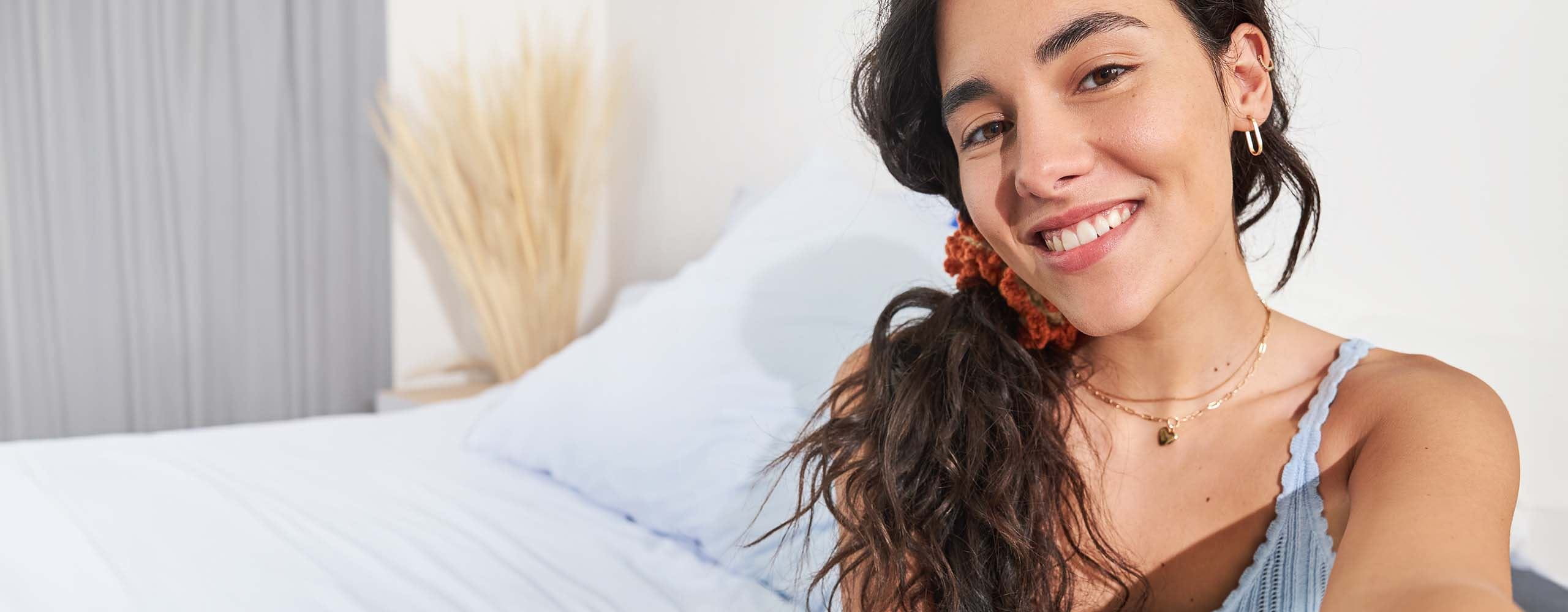 A person smiling while appearing to take a selfie photo while seated on a light coloured bed.