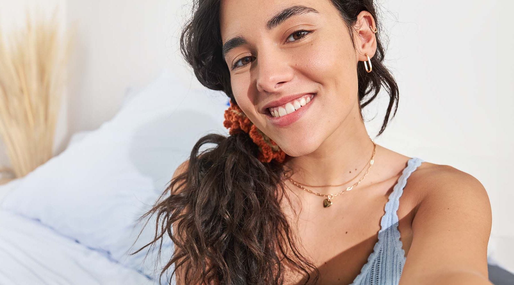 A person smiling while appearing to take a selfie photo while seated on a light coloured bed.