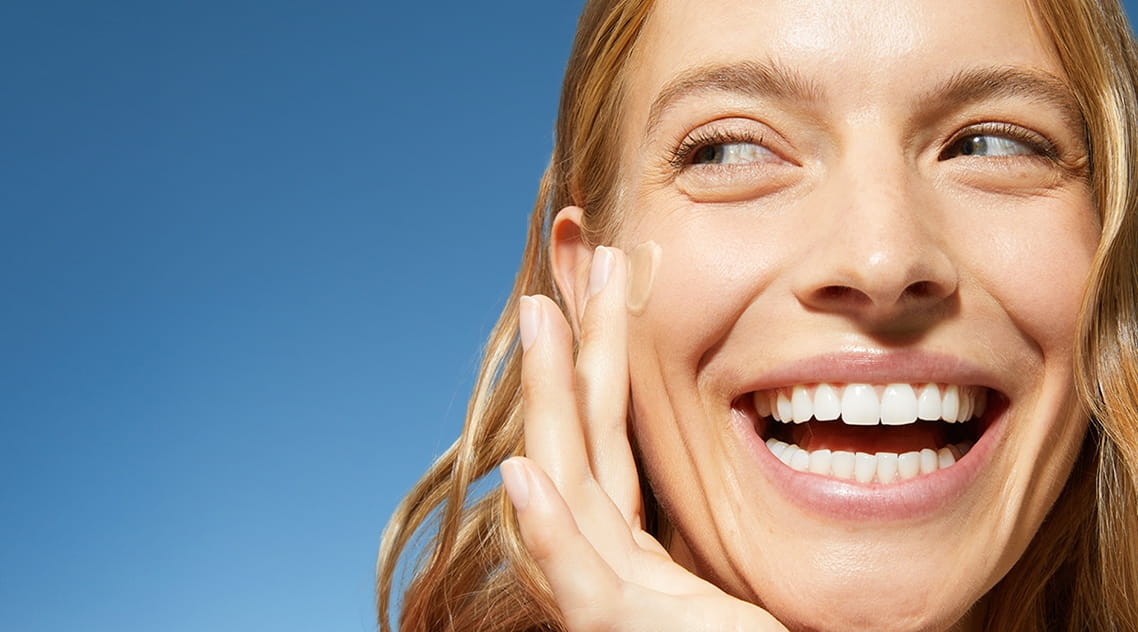 A woman smiles while swiping on daily fluid to her cheek.