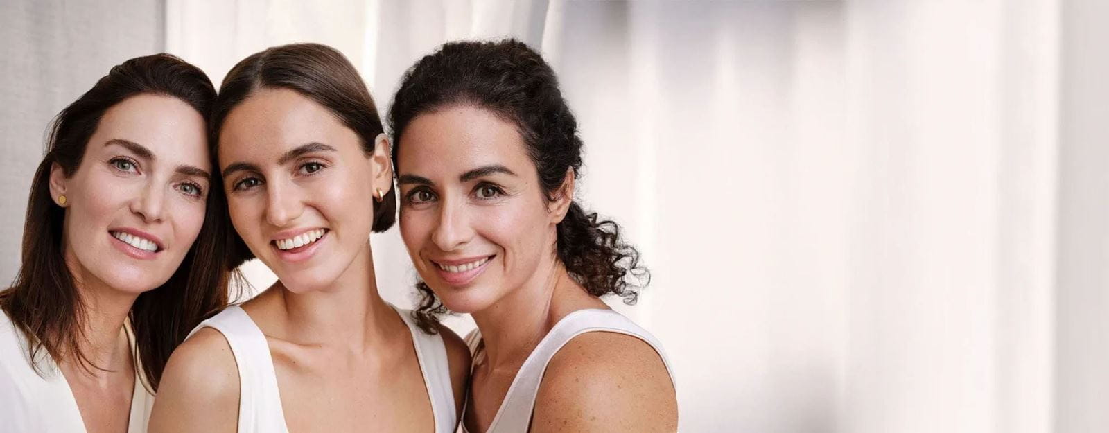 Trois femmes d'âge moyen sourient à la caméra, montrant une réduction des rides faciales, des taches brunes et des signes de vieillissement sur leur visage.