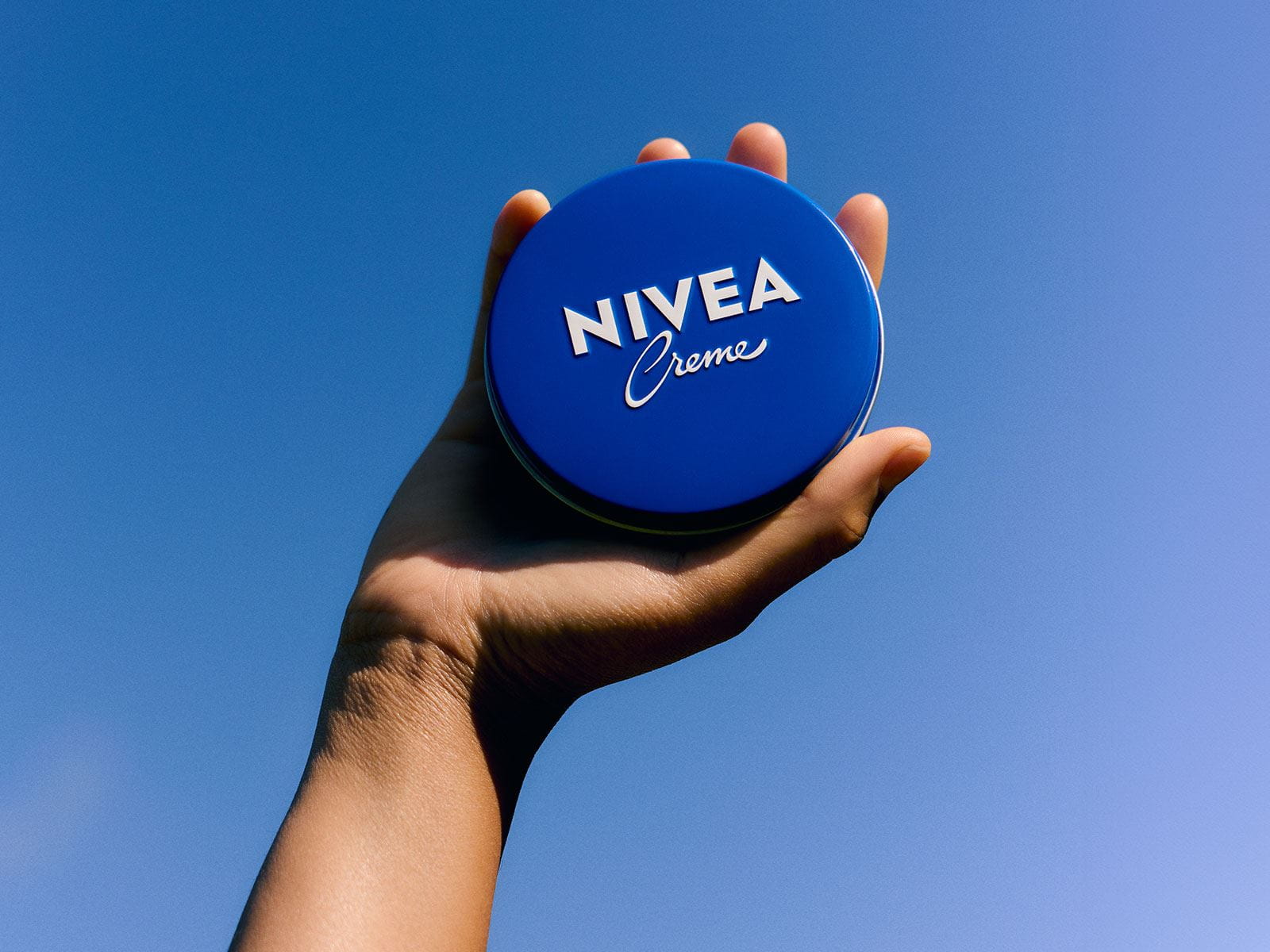 A NIVEA Creme blue tin being held up in the air, against a blue sky background.