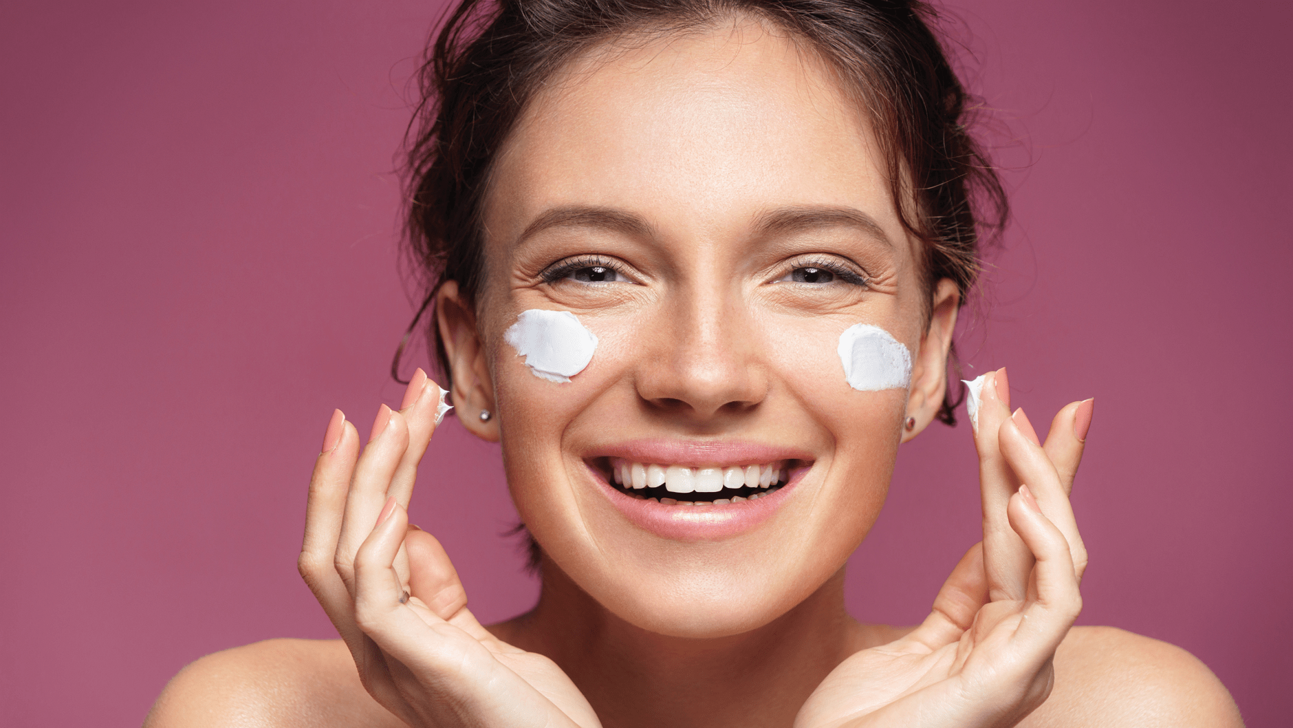 Smiling person applying cream to cheeks against pink background.