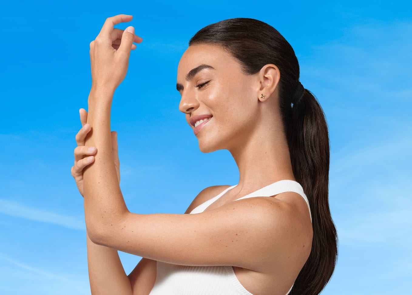 A woman in a white tank top smiling and applying lotion