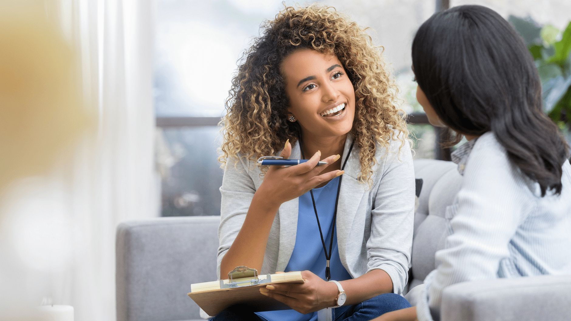 Two women having a conversation on a sofa, one holding a clipboard.