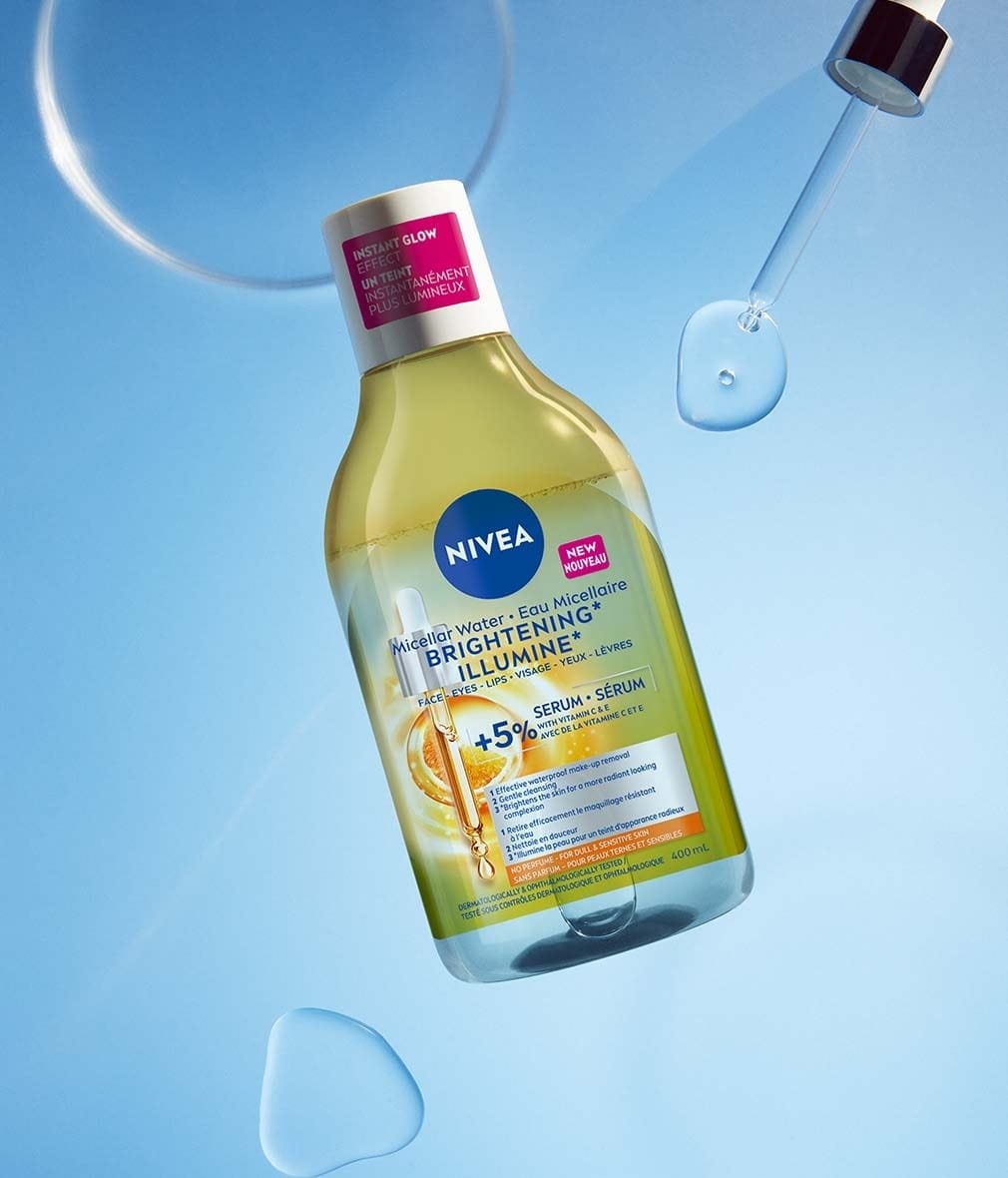 Un flacon d'Eau Micellaire NIVEA Illumine avec sérum est posé sur un fond bleu, entouré de gouttelettes d'eau. Le flacon contient de l'eau micellaire et l'étiquette mentionne la formule à 5 % de sérum avec vitamines C et E, ainsi qu'un teint instantanément plus lumineux. Un compte-gouttes laisse voir une goutte de sérum.