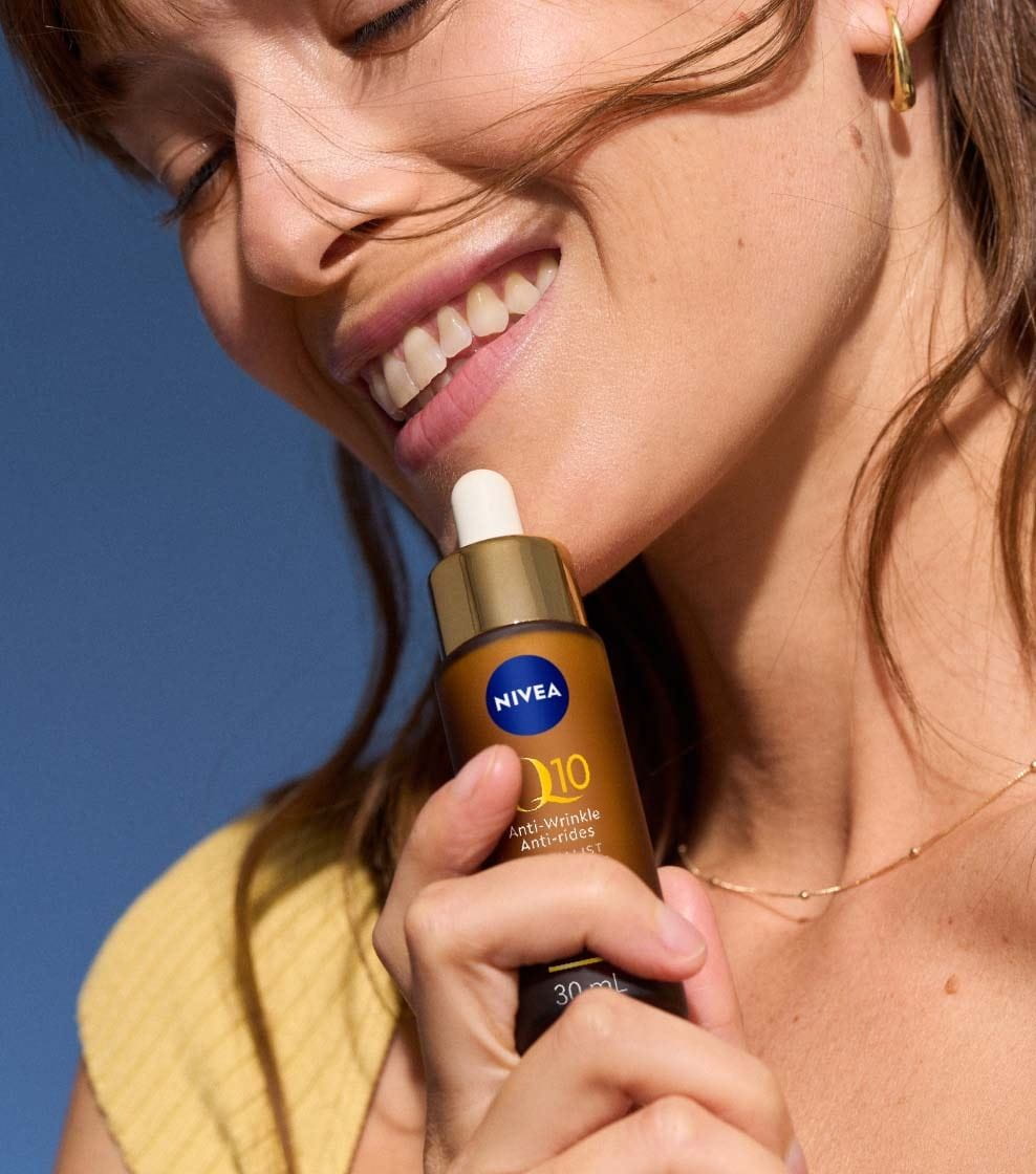 "A woman in a yellow top is smiling and holding a bottle of NIVEA Q10 ANTI-WRINKLE SPECIALIST DUAL ACTION SERUM by her lips. "