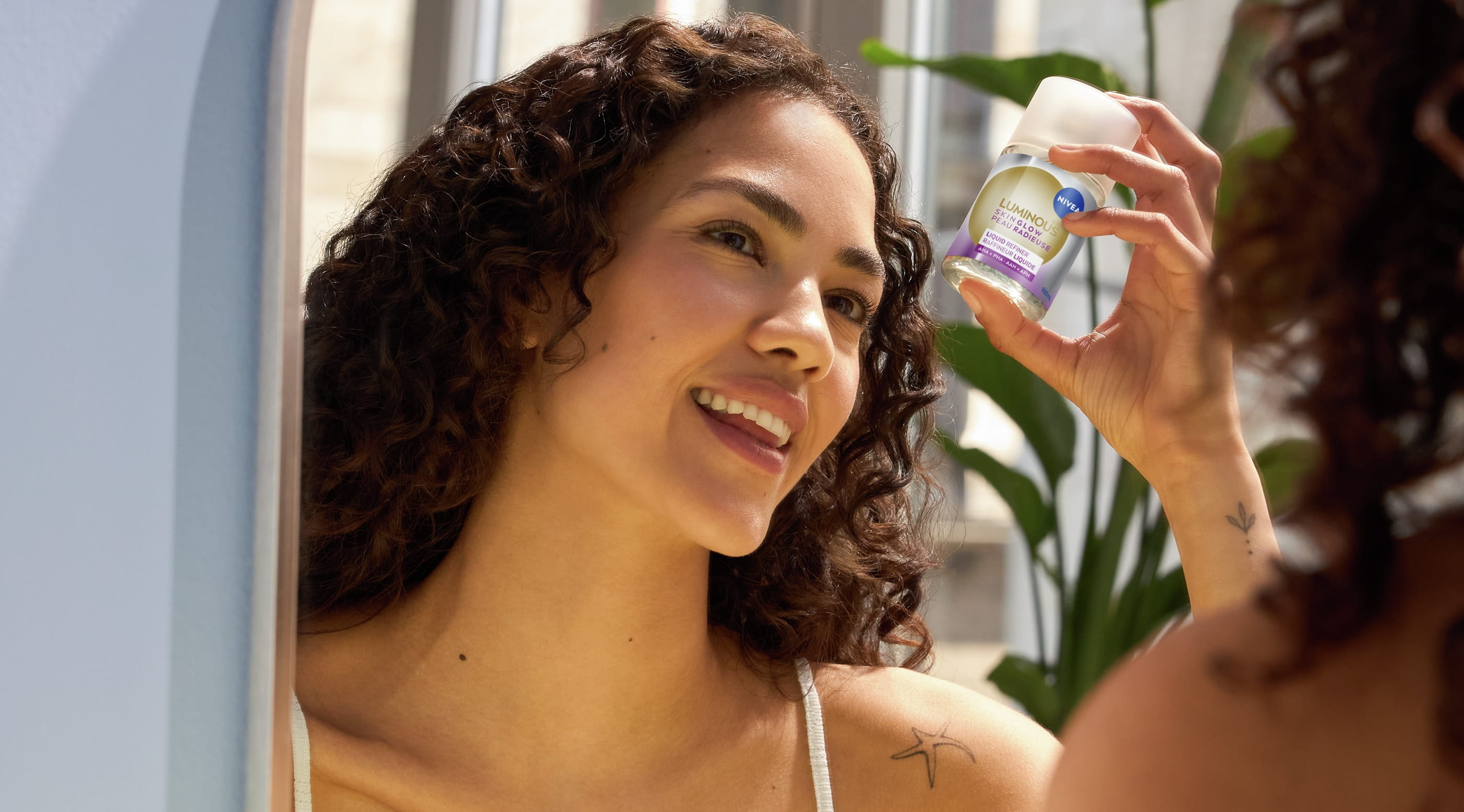 Person tittar på sig själv i spegeln, ler och håller upp NIVEA Skin Glow Daily Exfoliator