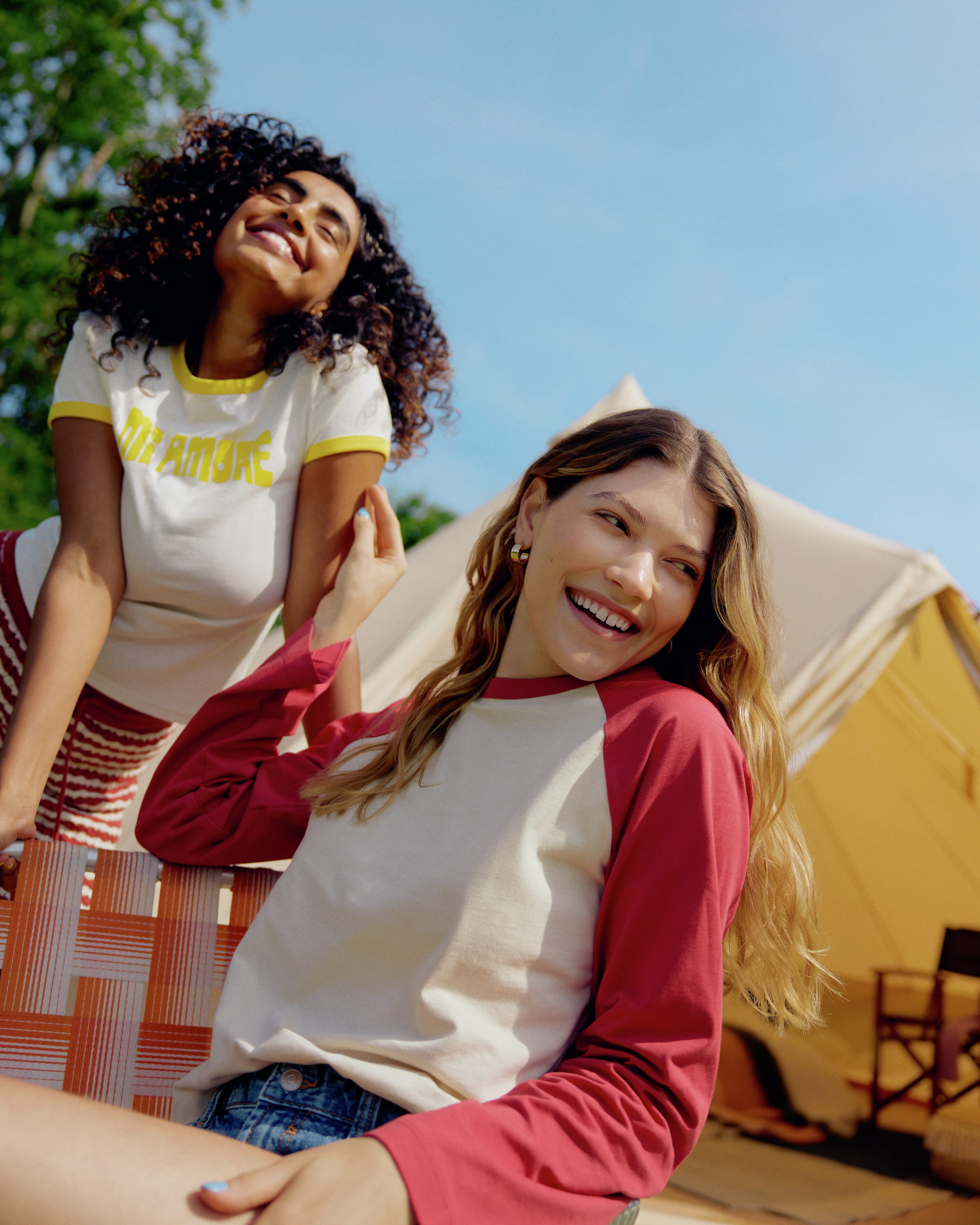 två kvinnor som skrattar, ena sitter på en stol och andra står bakom. Tält i bakgrunden