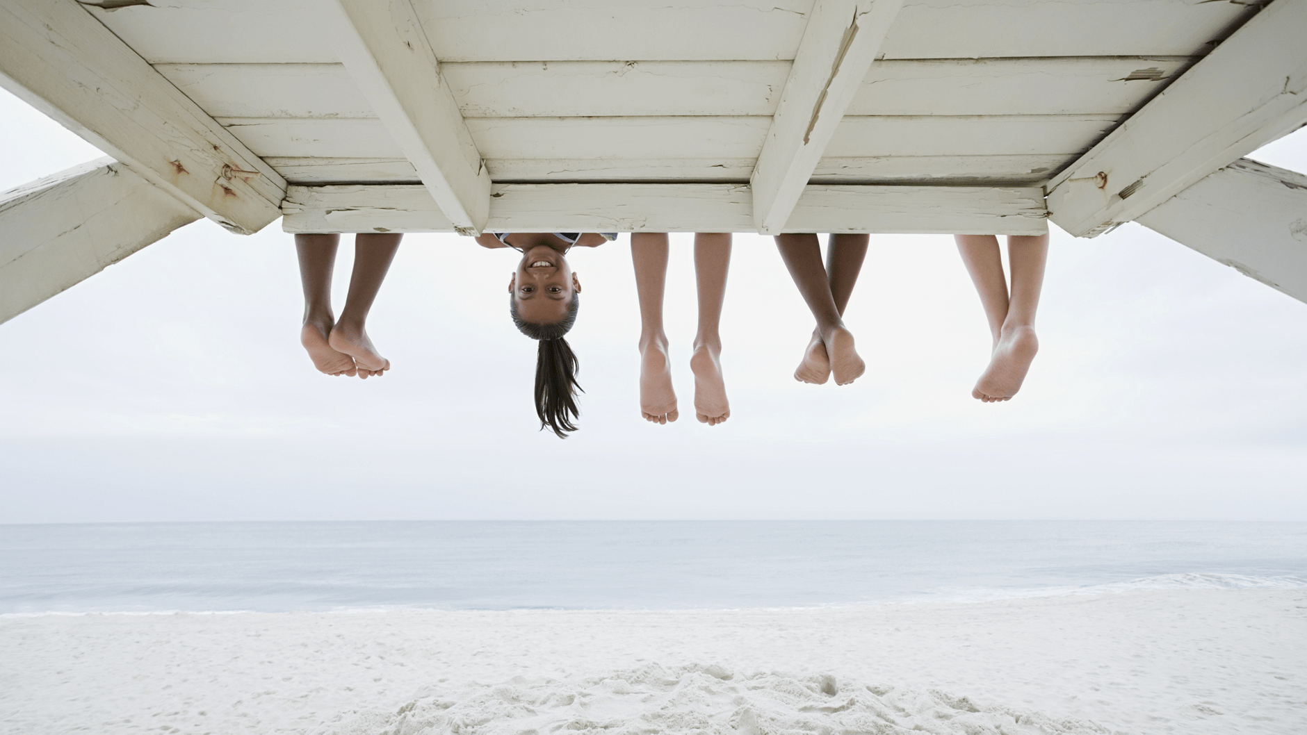 Children's feet and head hanging from a wooden structure at the beach.