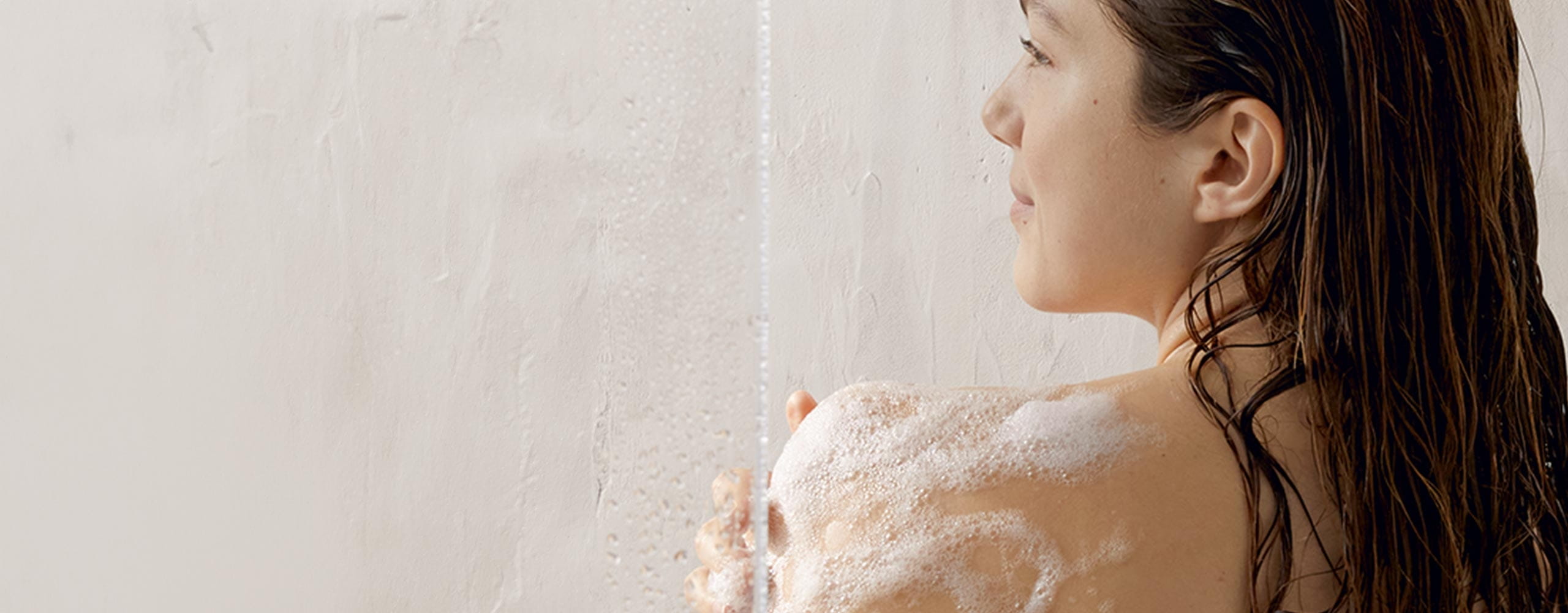 A back view of a person standing in a cream coloured shower with foaming soap seen on their body.