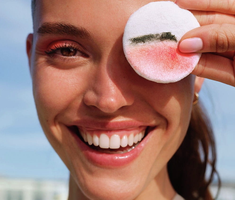 A young woman smiles laughingly and holds a cotton pad over her eye showing pink and black make-up residue.
