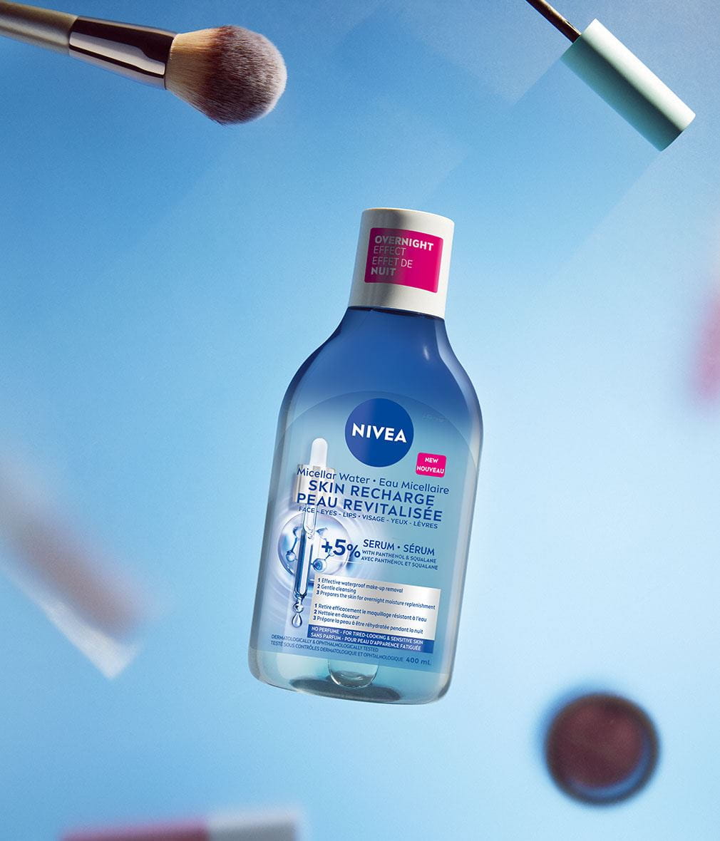 Un flacon d'Eau Micellaire NIVEA Peau revitalisée infusée au sérum est centré sur un fond bleu, entouré de brosses, pinceaux et cosmétiques flous à l'arrière-plan. L'étiquette du flacon indique l'effet de nuit du produit et sa formule à 5 % de sérum avec panthénol et squalane.