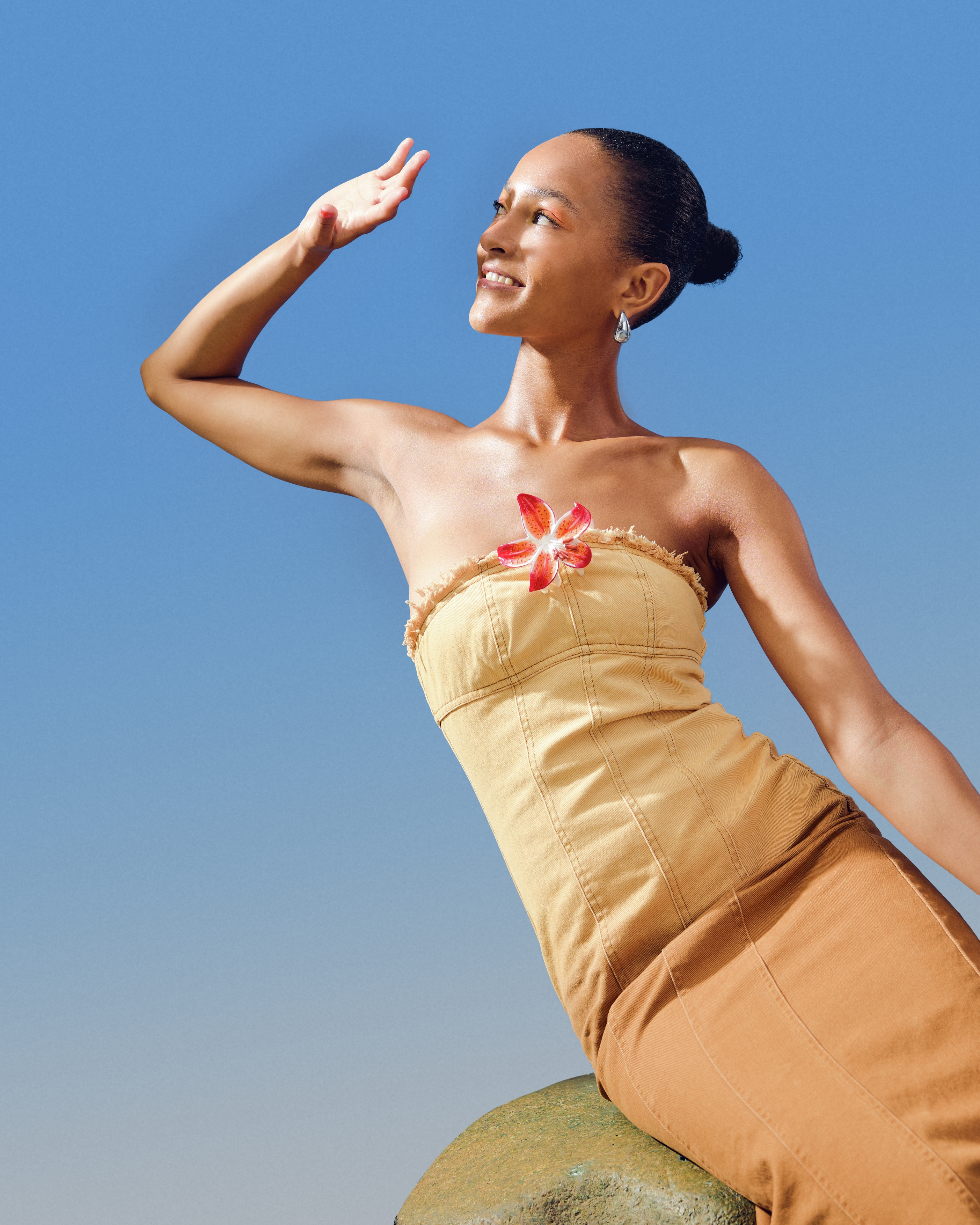 Tjej sitter på en sten, håller upp en hand och kisar mot solen