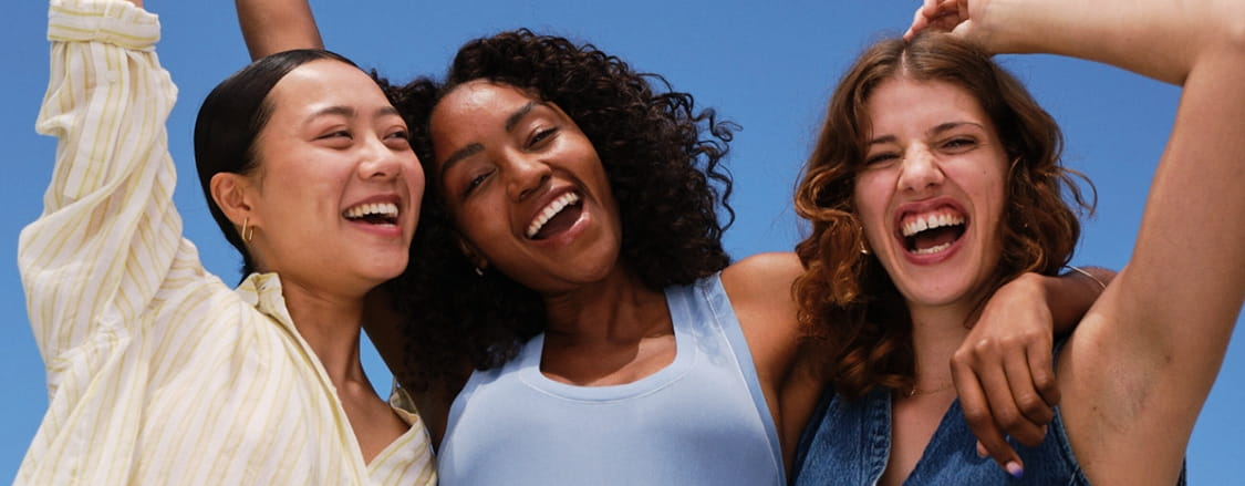 Three laughing young women standing close together, with their arms raised and around each other, exposing their underarms.