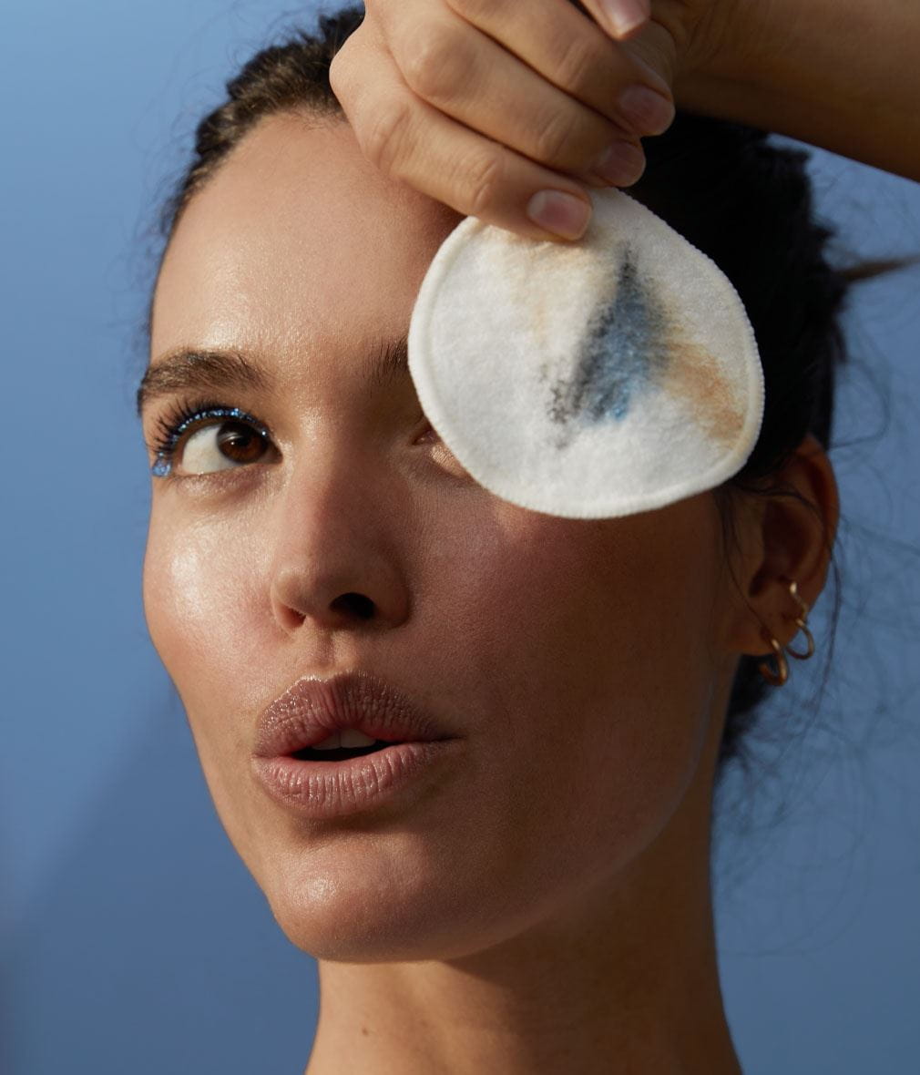 A woman holding a cotton pad with makeup residue against their eye, set against a blue background. Their facial expression appears focused and their skin looks dewy.