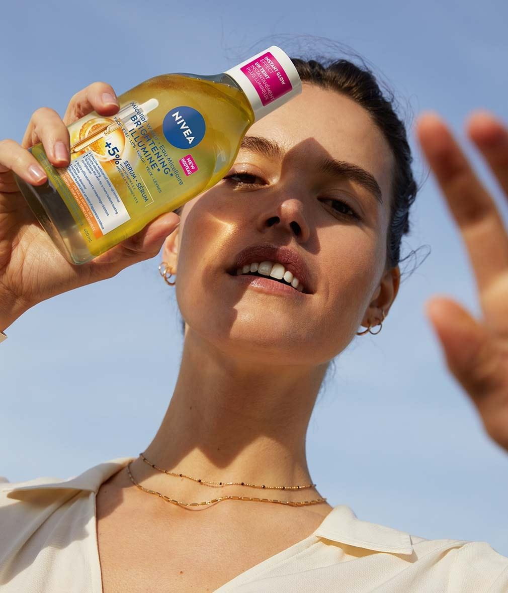 A woman holding a bottle of NIVEA Brightening Serum-Infused Micellar Water towards the camera with a sunny sky in the background. The bottle contains Micellar Water and the woman is smiling, wearing a light-colored top.