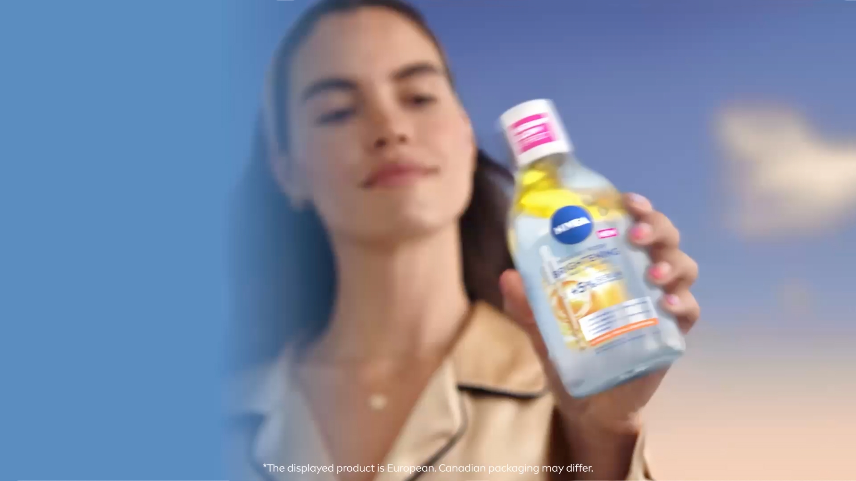 A woman wearing a silk top and a floral headband gently applies micellar water to her cheek. The background is a soft blue sky with clouds.