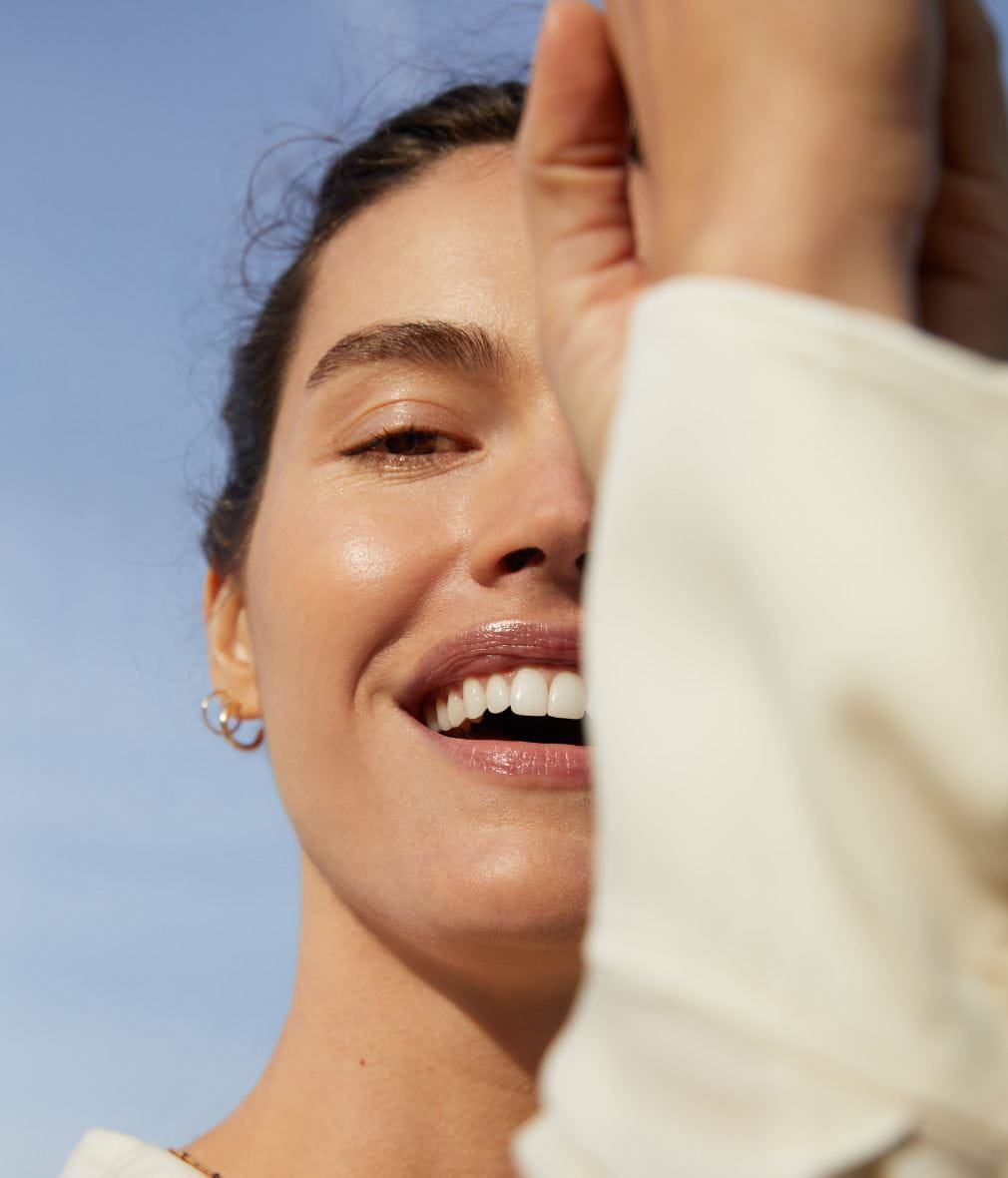 Photo d'une femme au grand sourire, sous un ciel bleu clair. À l'avant-plan, elle semble lever un bras vêtu d'une manche, ce qui obstrue partiellement le reste de l'image. L'expression de la femme dégage du bonheur.