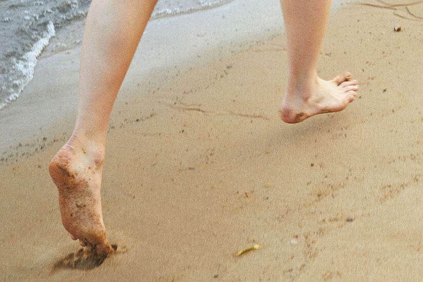 View of a model running barefoot on sand with water and waves crashing in the background.