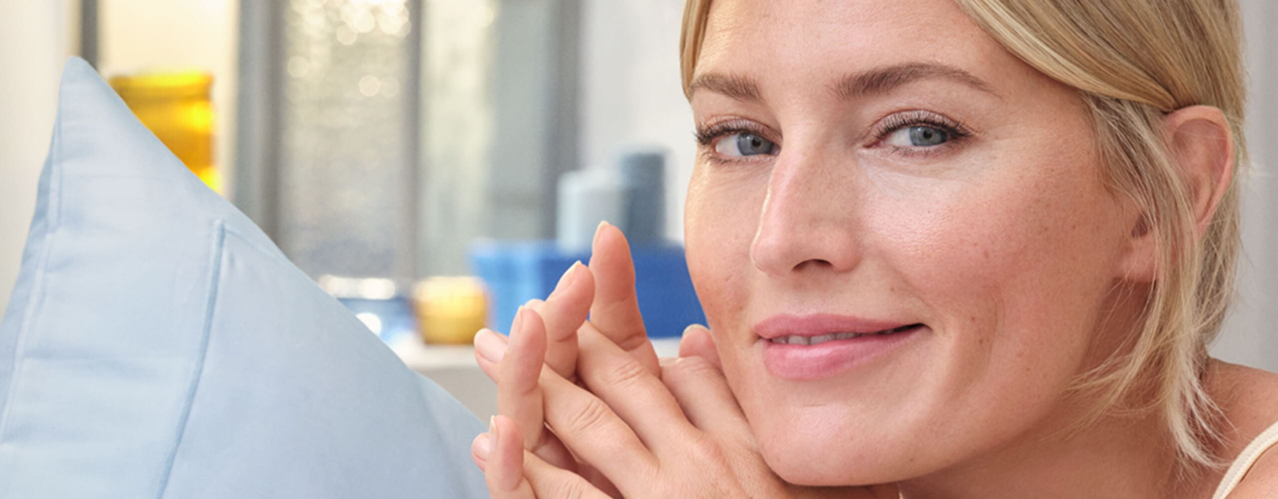 View of a female model with blonde hair and blue eyes smiling with light blue coloured pillows behind.
