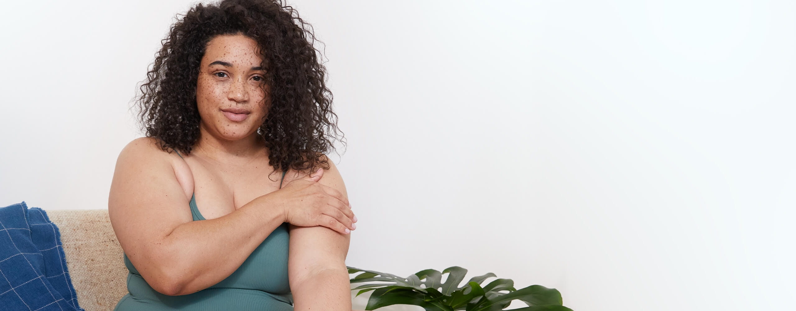 View of a female model with dark curly hair wearing a green coloured top smiling and holding her left arm.