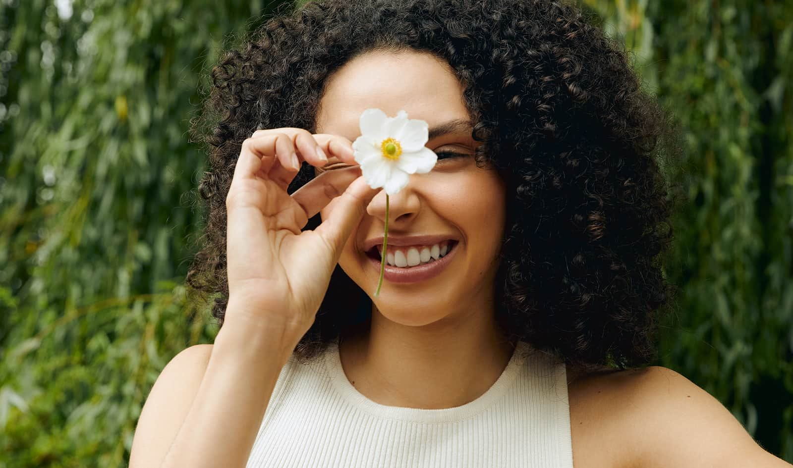 View of a female model with dark curly hair holding a white flower in front of her face with trees in the background.