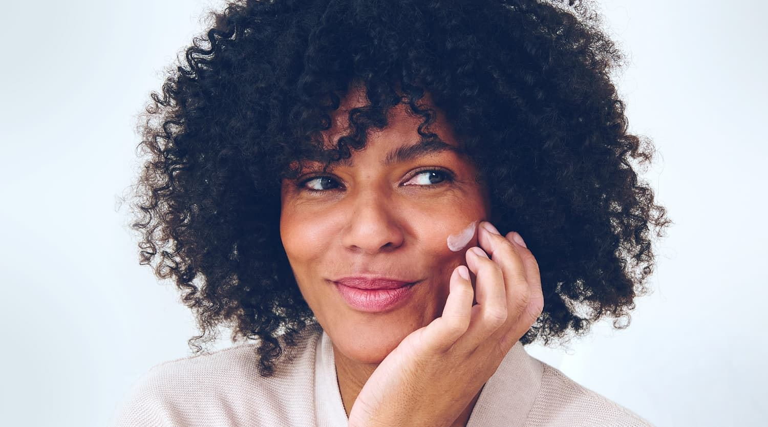 Front view of a female model with dark curly hair smiling with a smudge of NIVEA cream on her left cheek.