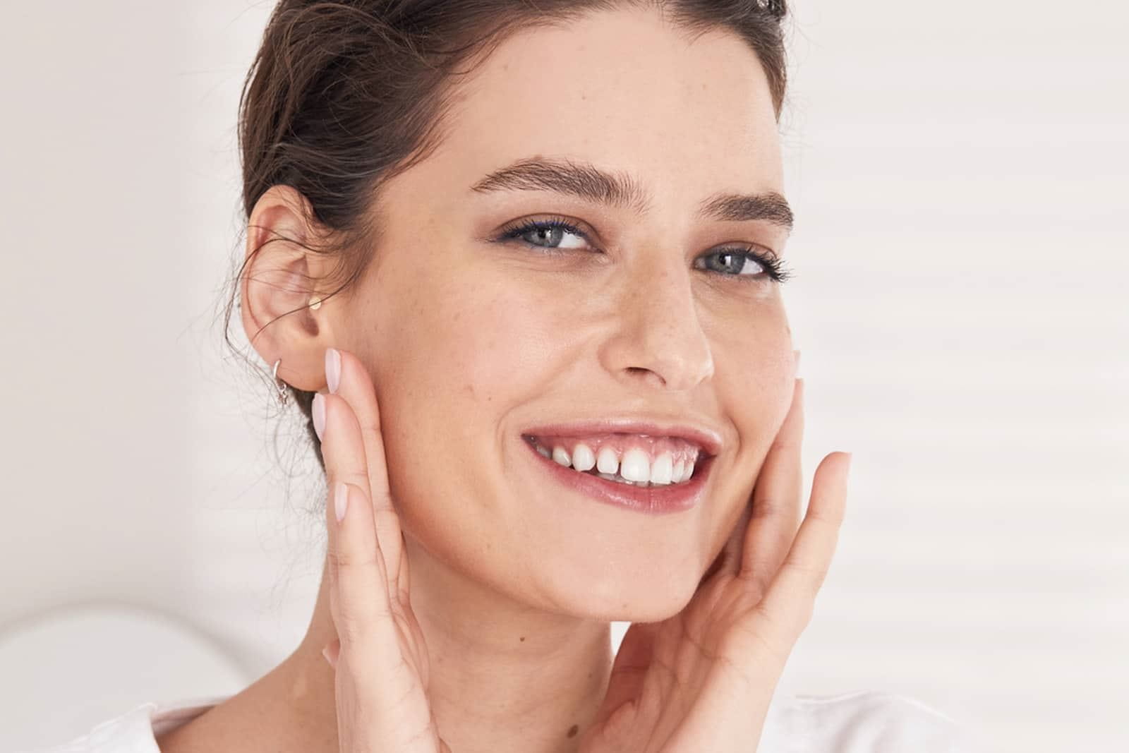 View of a female model with brown hair and blue eyes smiling while touching her face against a white background.