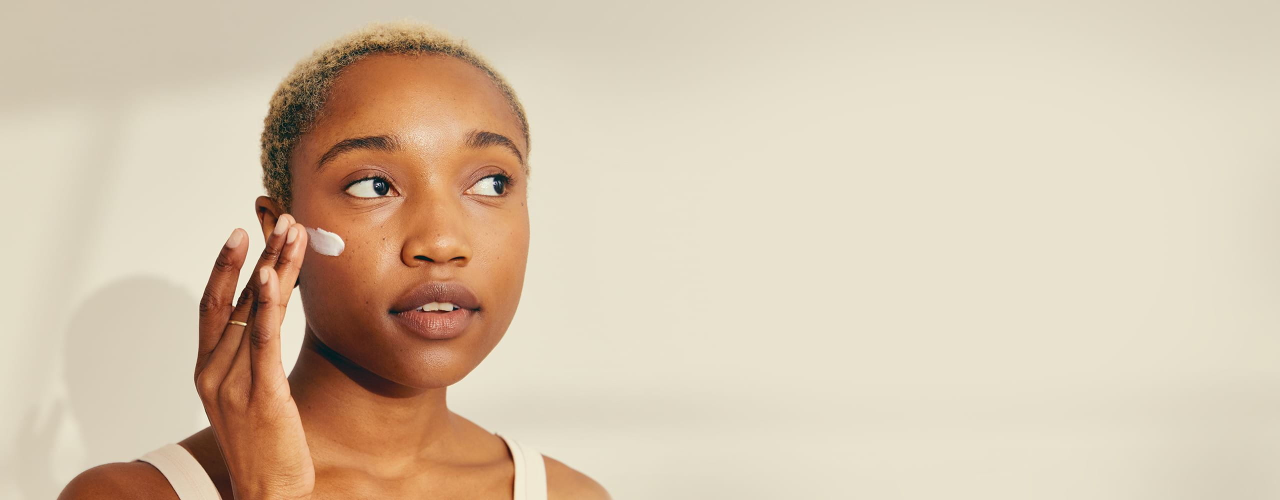 Mannequin féminin aux cheveux blonds courts appliquant de la crème NIVEA sur sa joue droite, sur fond beige.