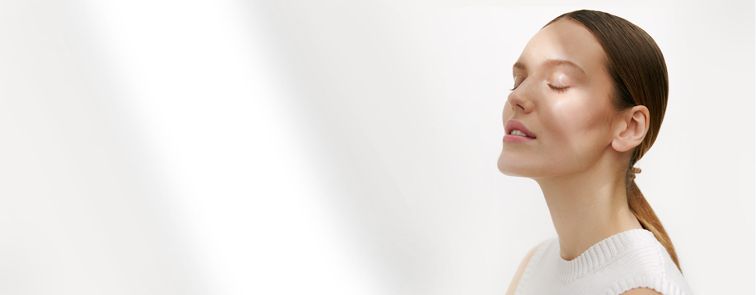 View of model wearing a white knit top with eyes closed and shadows appearing on face against a white background.