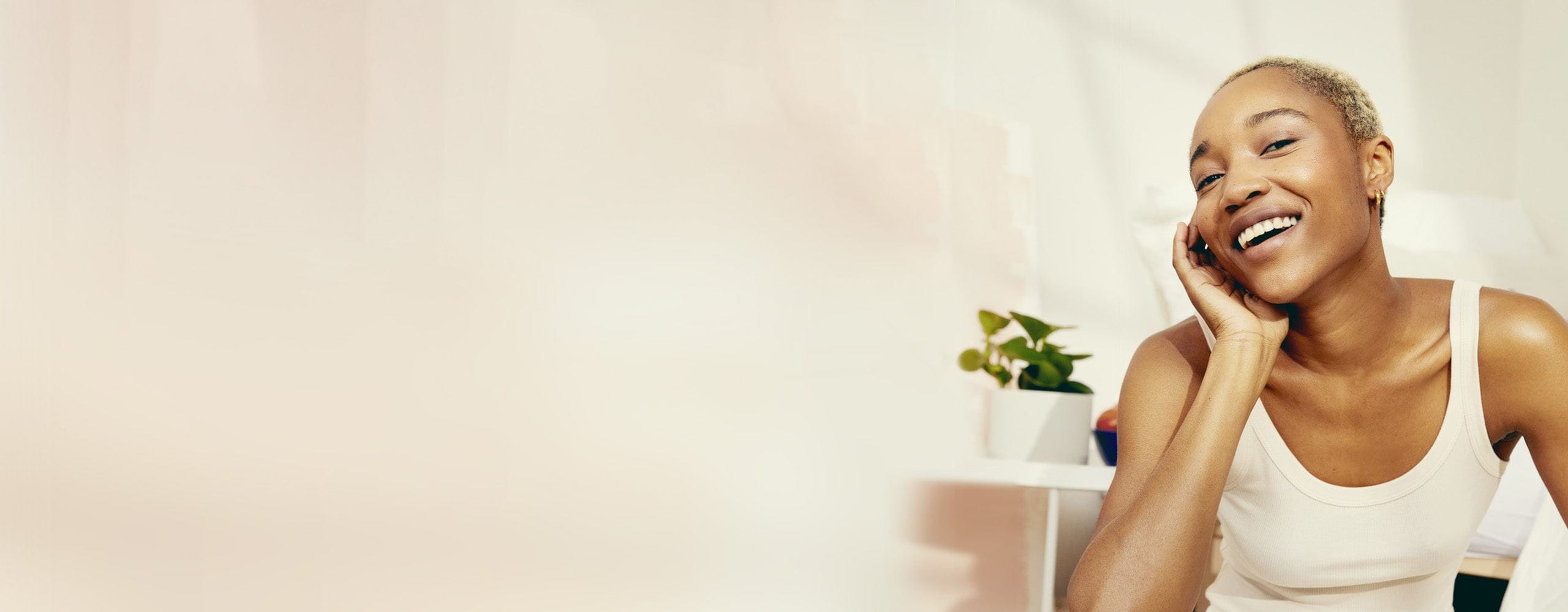 View of a smiling model wearing a white tank top with right hand on right side of face with a green plant in the background.