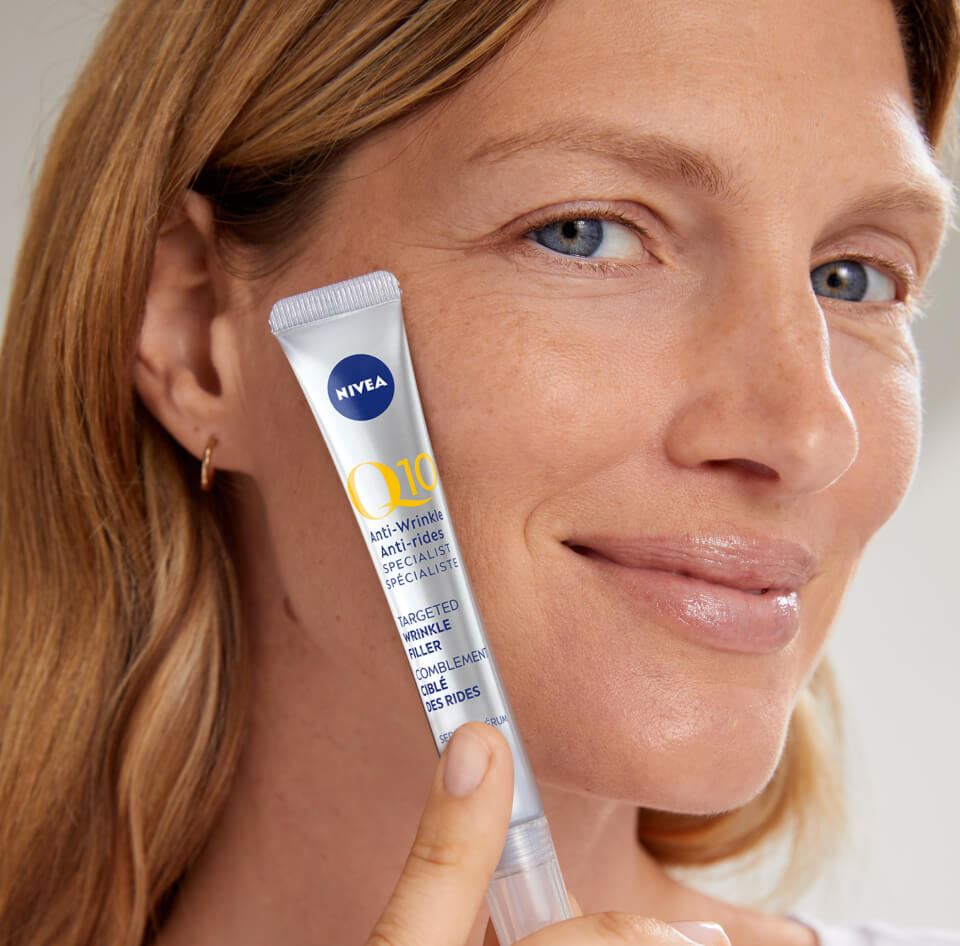 Vue d’un mannequin féminin souriant tout en tenant le produit NIVEA Q10 Anti-rides Spécialiste Comblement ciblé des rides contre sa joue.