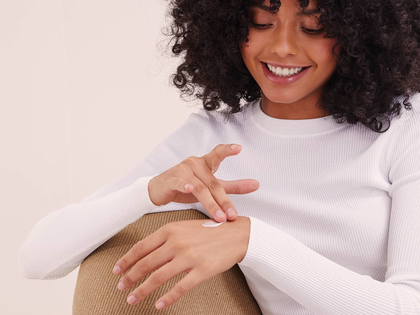 A person smiling and looking down at their hand while applying a cream product.  