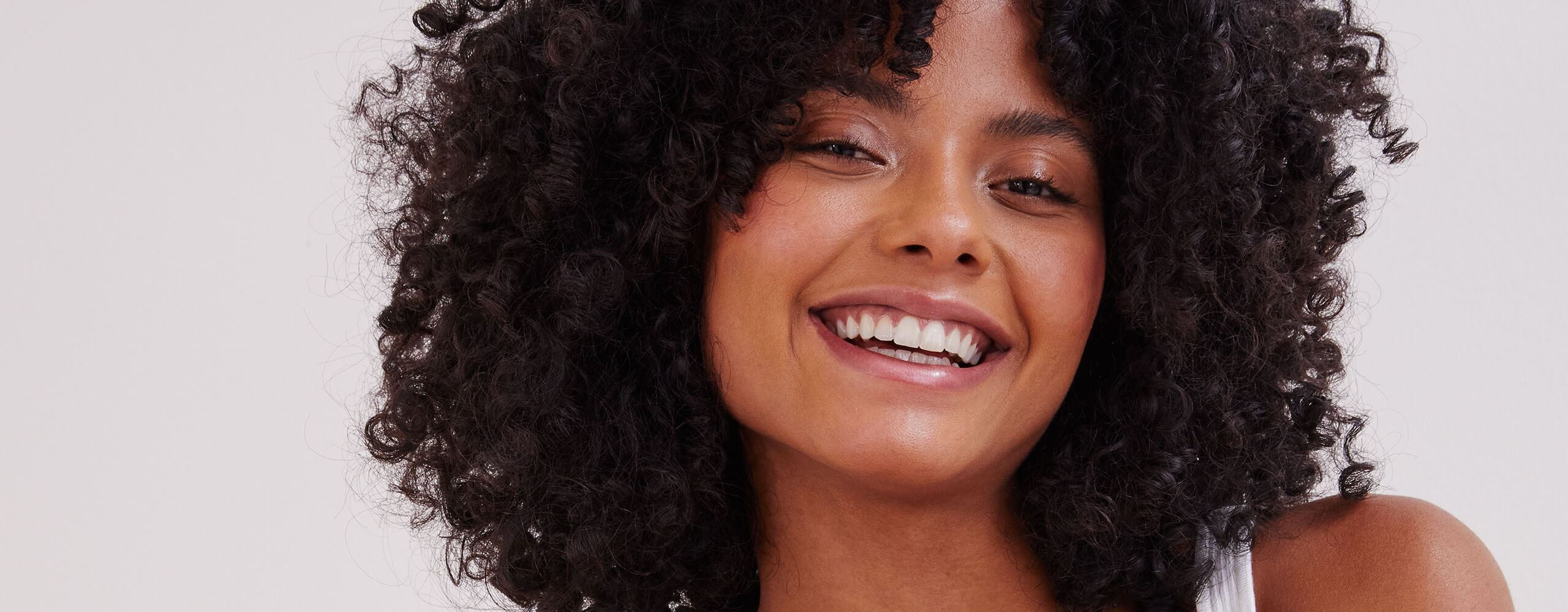 Vue d'une personne aux cheveux courts et bouclés de couleur foncée souriant sur un fond blanc.