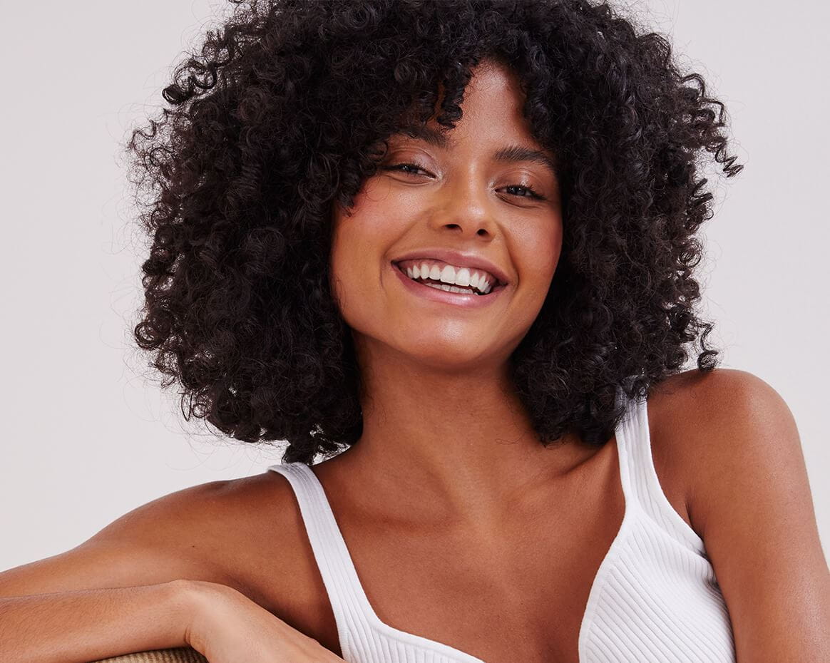 A view of a person with dark-coloured short, curly hair smiling against a white-coloured background.