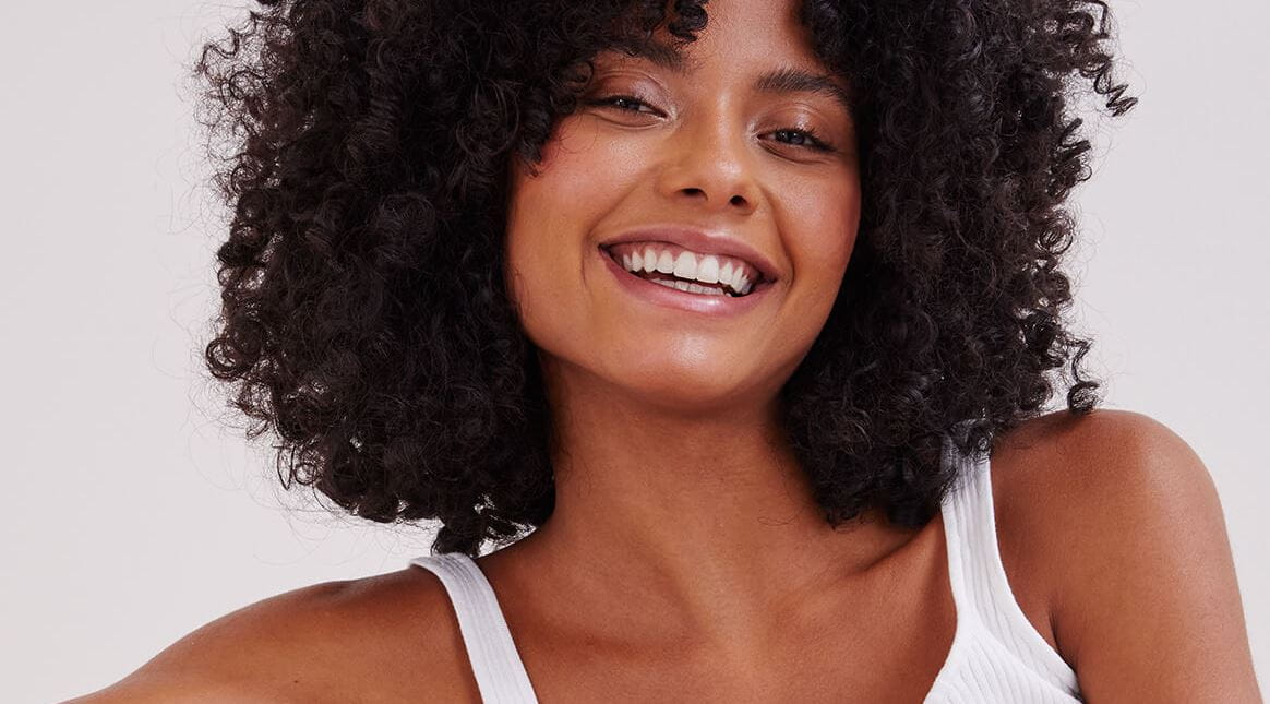 Vue d'une personne aux cheveux courts et bouclés de couleur foncée souriant sur un fond blanc.