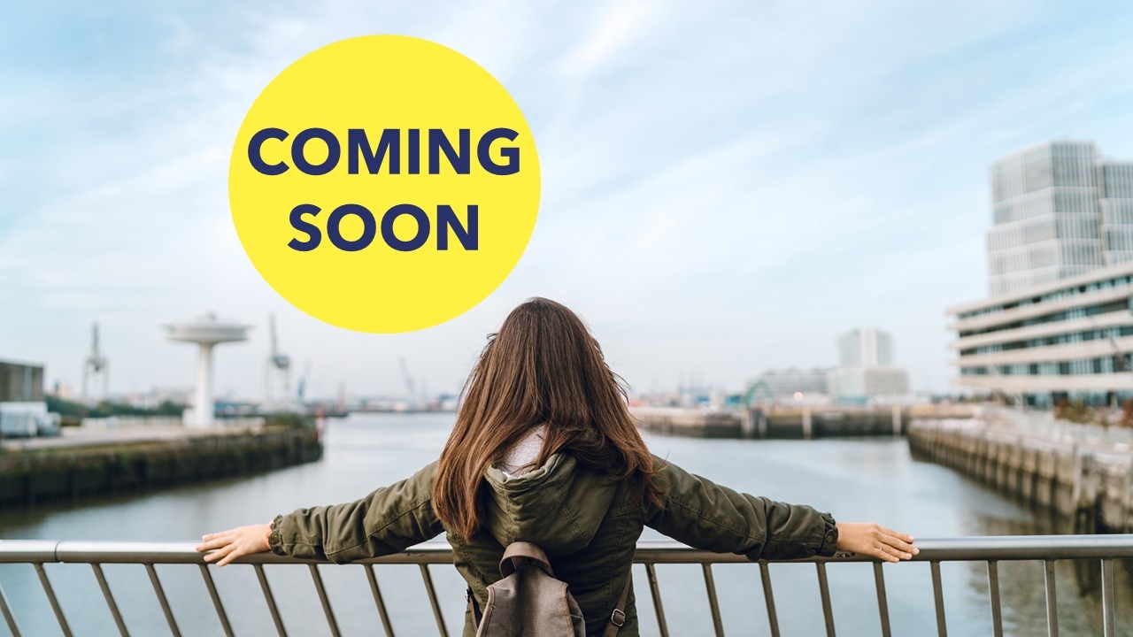 Person standing by a river, facing away, with cityscape in background.